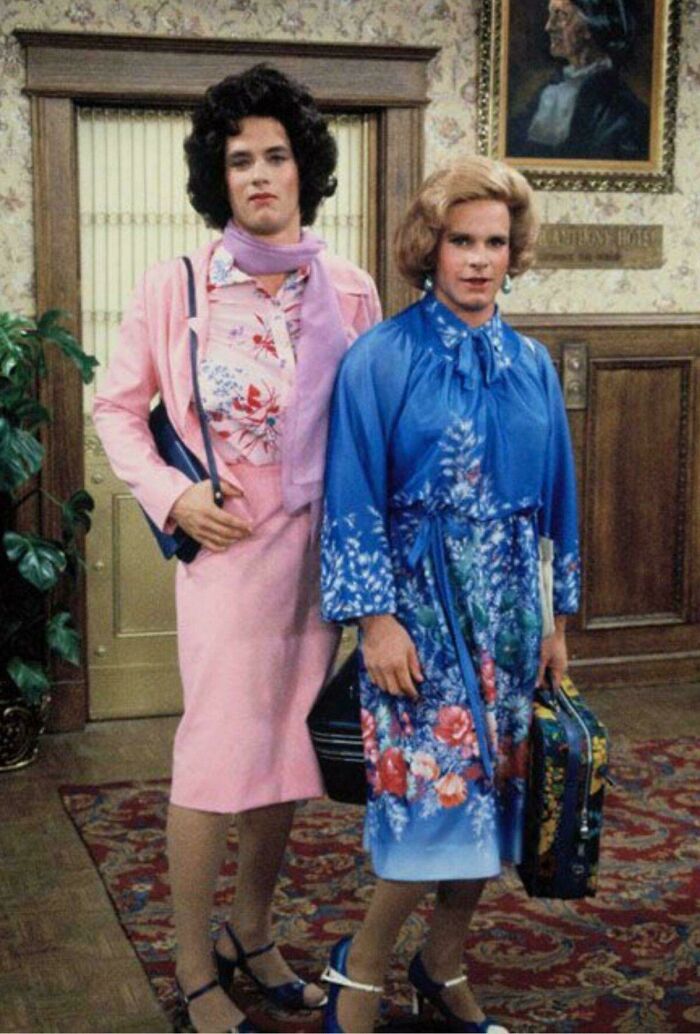 Two men dressed in vintage floral dresses and wigs standing indoors, a nostalgic image that makes people feel very old.