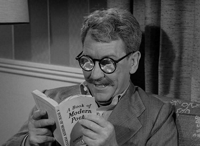 Black and white image of a man with glasses smiling while reading a vintage book, evoking nostalgia and feeling old.