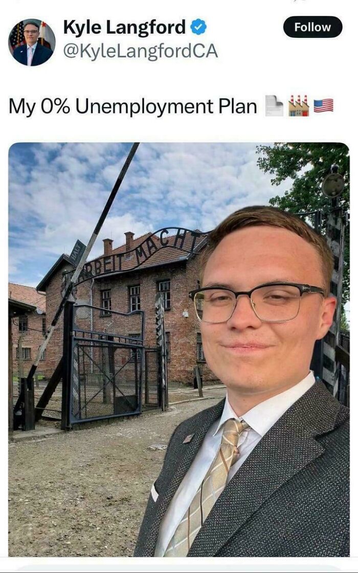 Young man in glasses and suit smiling for selfie in front of Auschwitz gate, illustrating boring dystopia in modern life.