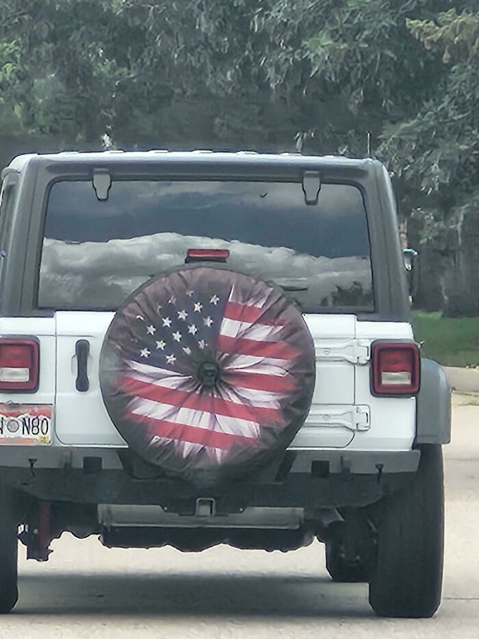 White SUV with a crumpled American flag tire cover showcasing a failed attempt at patriotic decoration on a sunny day.