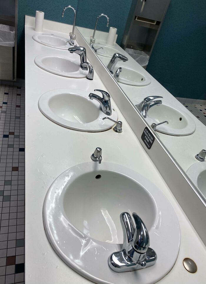 Public restroom sinks with misaligned faucets and a mirror reflecting imperfect bathroom fixtures, highlighting OCD annoyances.