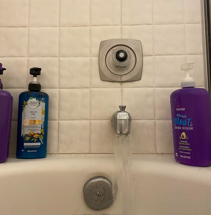 Bathtub faucet with water flowing unevenly, surrounded by shampoo and conditioner bottles, highlighting imperfect bathroom details.