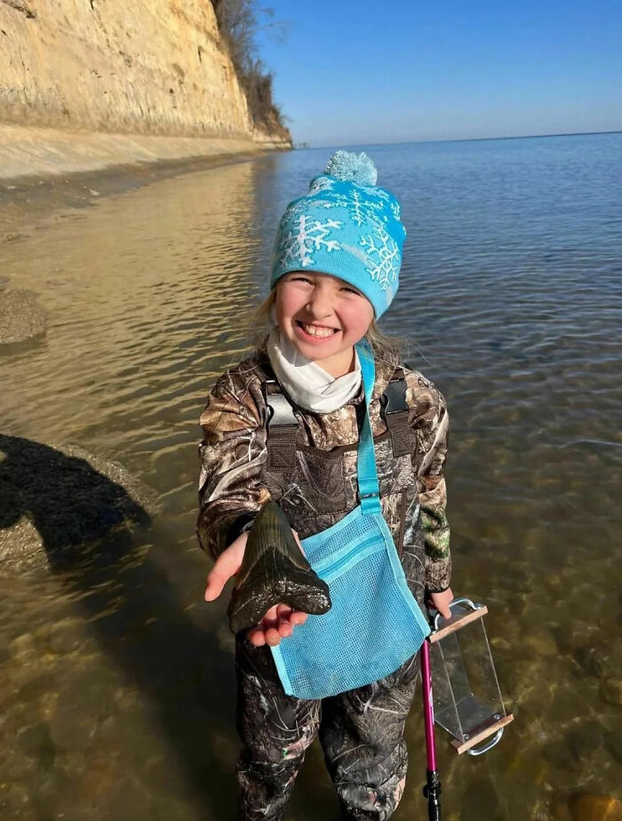 Young girl in outdoor gear smiling and holding a large shark tooth near water, featured in an online group sharing interesting facts.