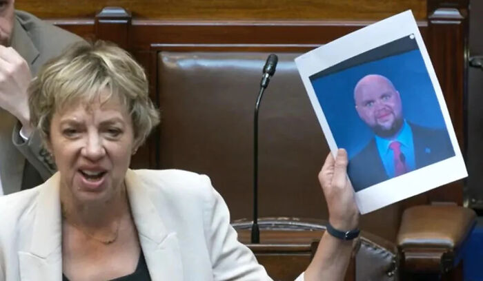 Woman in a white blazer holding a printed photo during a discussion in a formal meeting room setting.