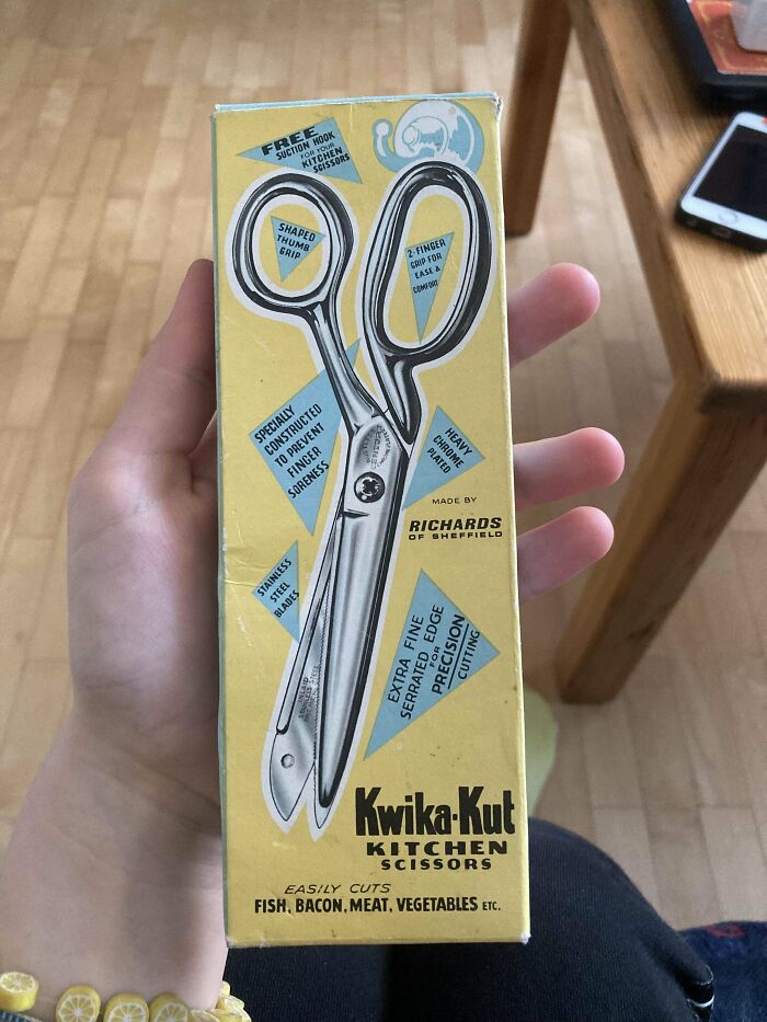 Hand holding vintage 1950s kitchen scissors box with retro design and product details on wooden floor background.