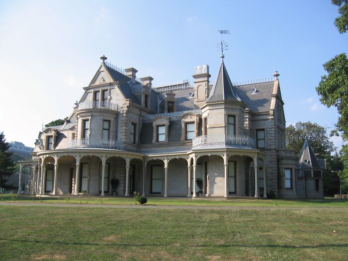 Large historic home with Victorian architectural features and wraparound porch set in a grassy American heritage landscape.