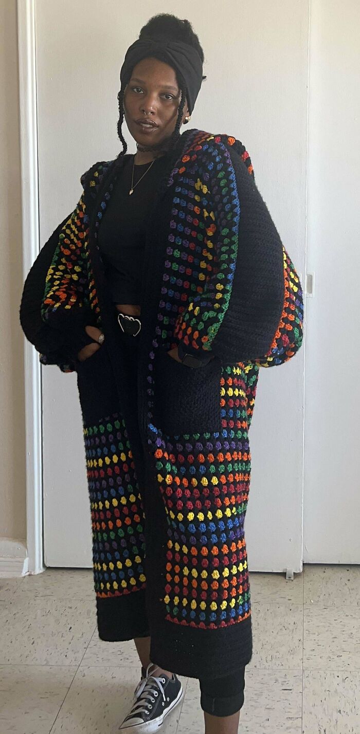 Person wearing a colorful crochet cardigan with vibrant rainbow patterns standing indoors against a plain wall.