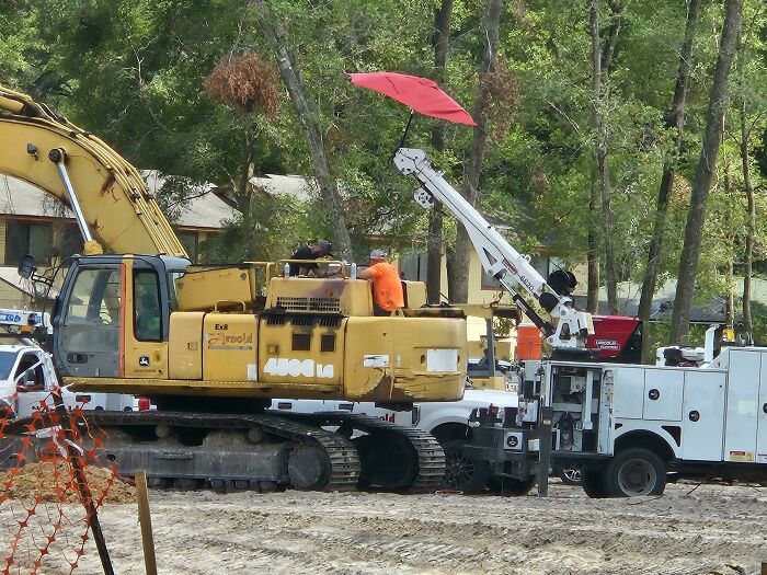 Construction workers on heavy machinery using a red umbrella for shade during heatwaves in a dusty outdoor site.