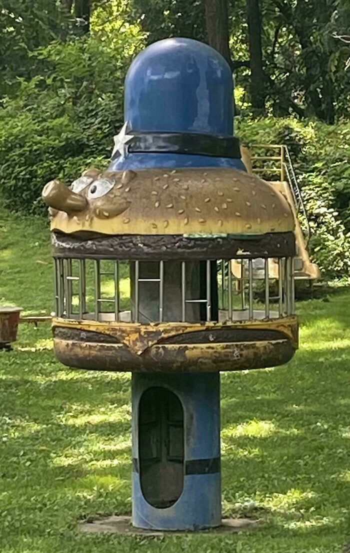 Old nostalgic playground ride shaped like a whimsical burger with a blue hat in a grassy park area.