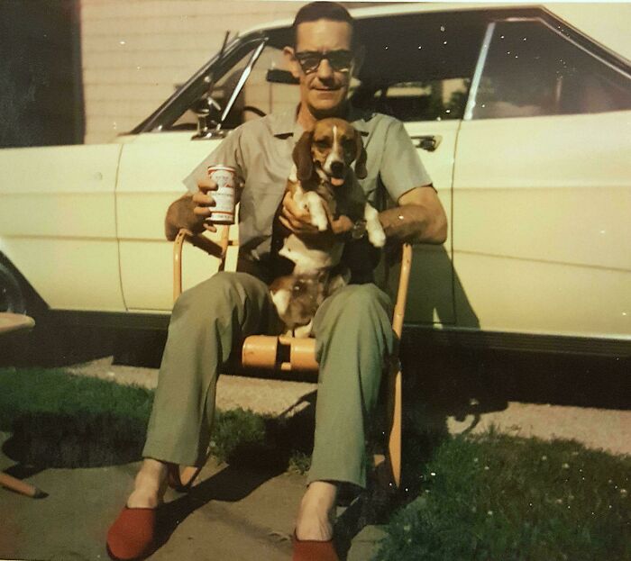 Man sitting outside a vintage car holding a dog and a beverage, a candid glimpse into the past captured in a casual moment.
