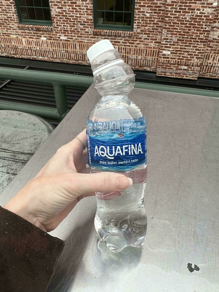 Hand holding a severely deformed Aquafina water bottle warped by the extreme heatwave outdoors on a metal surface.