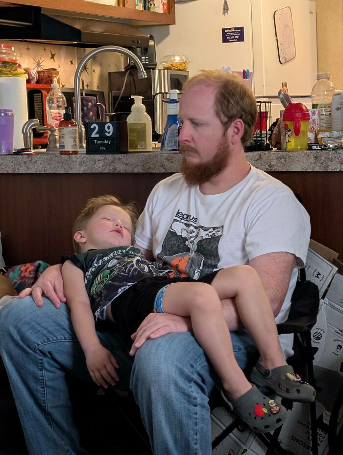 A father holding his sleeping child in a kitchen, an accidental Renaissance photo capturing a quiet, tender moment.