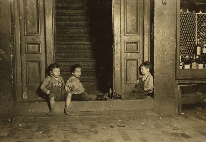 Three boys in worn clothes sit on a stoop in a somber historic photo telling a gut-wrenching story.