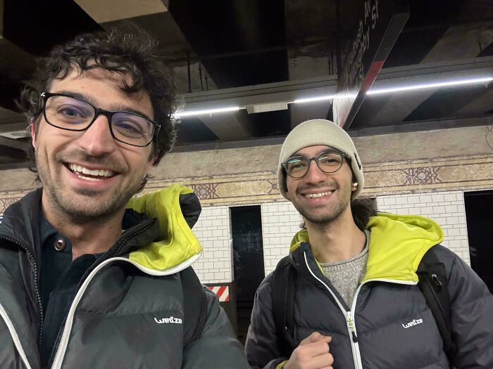 Two men wearing glasses and similar jackets, smiling in a subway station, showcasing rare coincidences in photos.