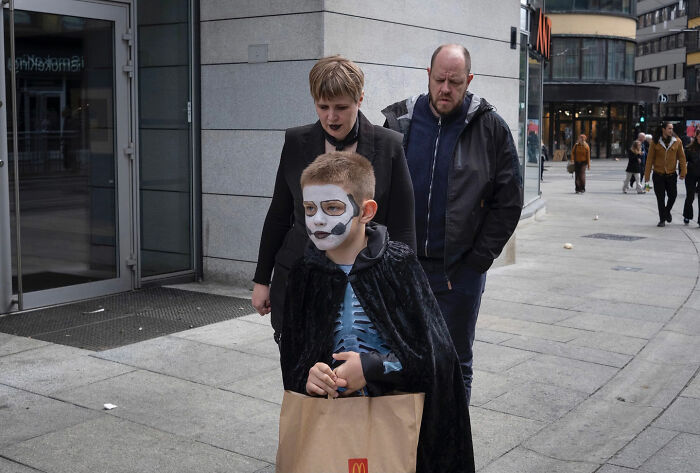 Child in skeleton costume and face paint holding a McDonald's bag, followed by two adults on a city sidewalk, street photography.