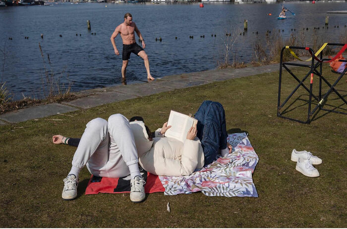 Couple lying on blankets by the water while a man prepares to jump in the background, a funny accidental image.