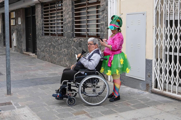 Elderly man in wheelchair accompanied by person wearing colorful outfit and funny hat on city sidewalk, street photography moment