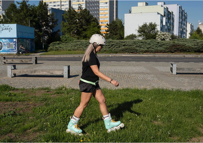 Woman wearing rollerblades and a helmet, walking on grass in an urban area, captured in a funny accidental street photography moment