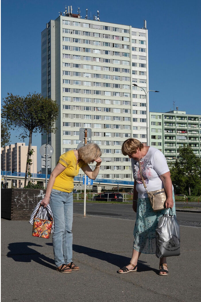 Two women looking down at the pavement in front of tall buildings, capturing a funny accidental moment by street photographer.