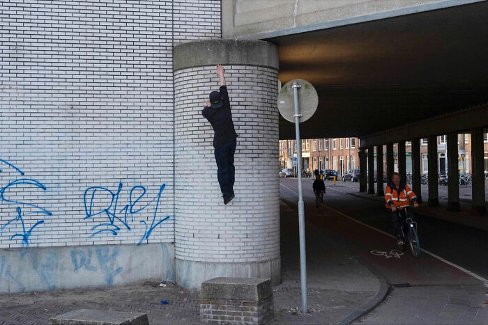 Person appearing to hang from a street pillar in funny accidental image taken at the right time by street photographer.
