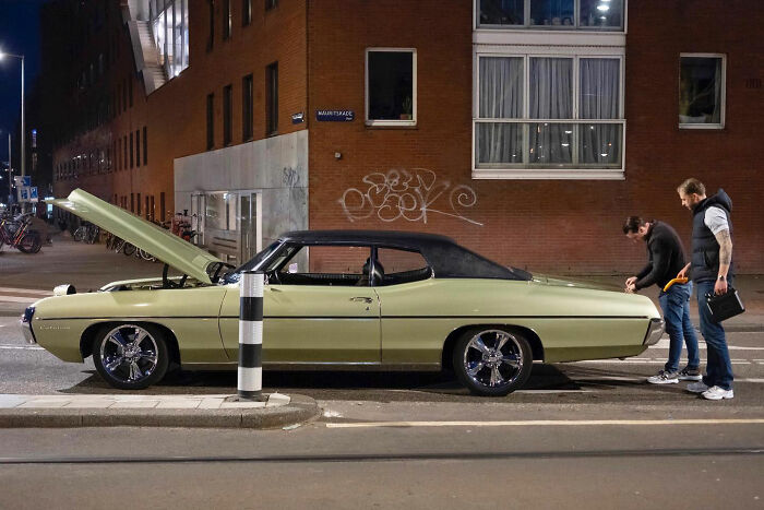 Classic car with open hood parked on street at night while two men inspect it in a funny accidental image by street photographer