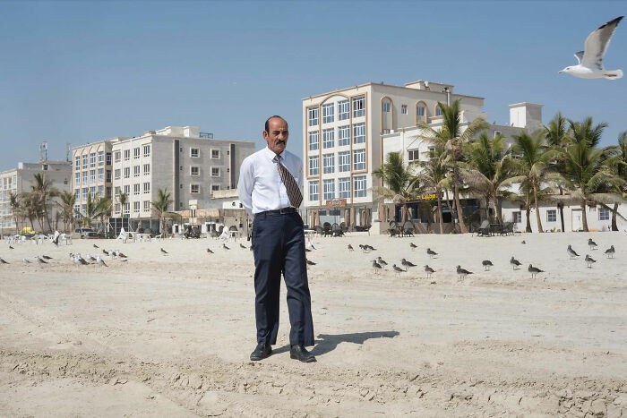 Man in formal attire standing on beach with seagulls and buildings, captured by street photographer at right time.