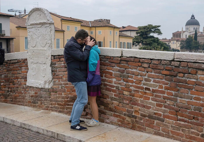 Couple sharing a kiss against a brick wall, captured as a funny accidental image by a street photographer.