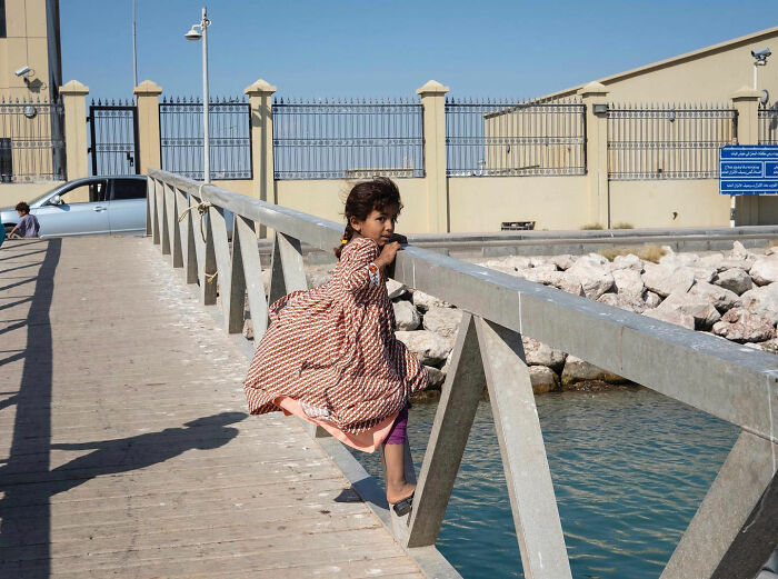 Young girl in a patterned dress sitting on a railing by the water, captured in a funny accidental street photographer image.
