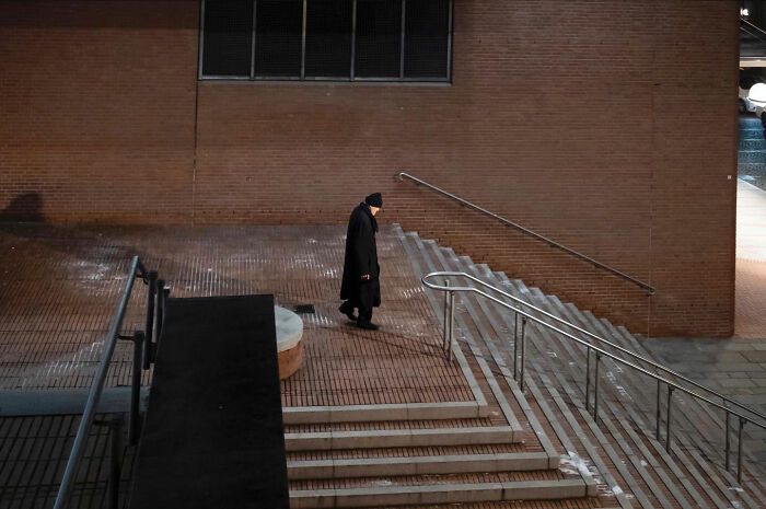 Person in black coat and hat walking by urban stairs, captured as a funny accidental image by street photographer.