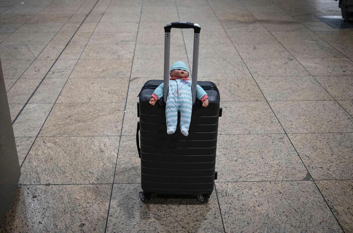 Doll dressed in striped pajamas positioned on black suitcase handle in street photographer's funny accidental image.