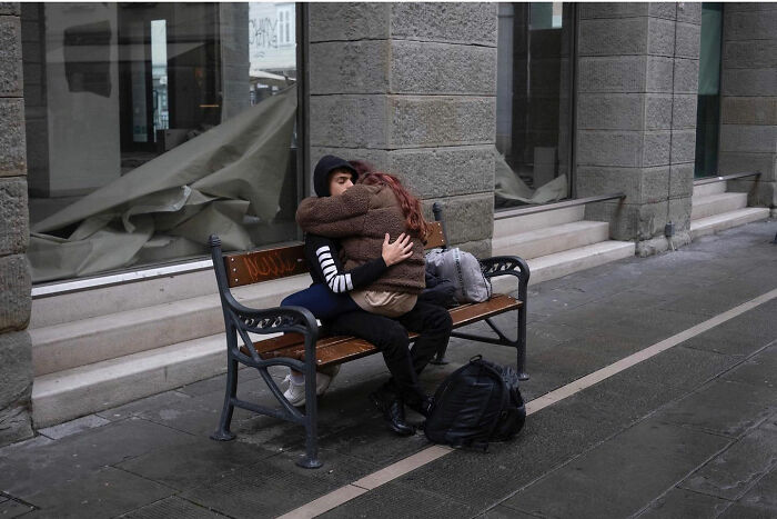 Couple embracing on a bench on a city sidewalk, captured in a funny accidental image by street photographer.