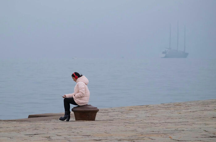 Woman in a pink jacket and red headphones sitting by the water, captured in a funny accidental image by street photographer.