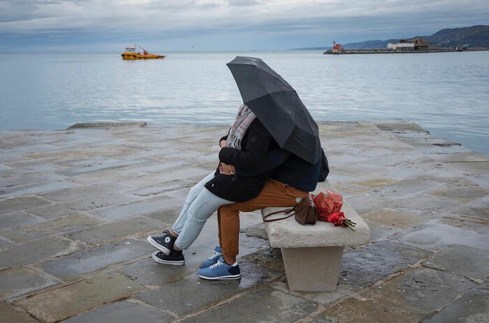 Couple sitting on a bench under an umbrella by the sea, a funny accidental image taken at the right time by street photographer.