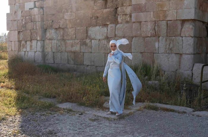 Woman in a flowing dress and headscarf walking by an old stone wall, captured in a funny accidental street photograph.