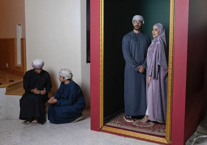 Two men sitting and talking next to a large ornate frame with a couple posing inside in traditional clothing.