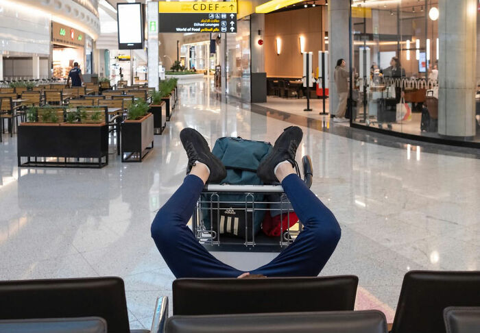 Person lying with legs up on luggage cart in airport corridor, a funny accidental street photography moment.