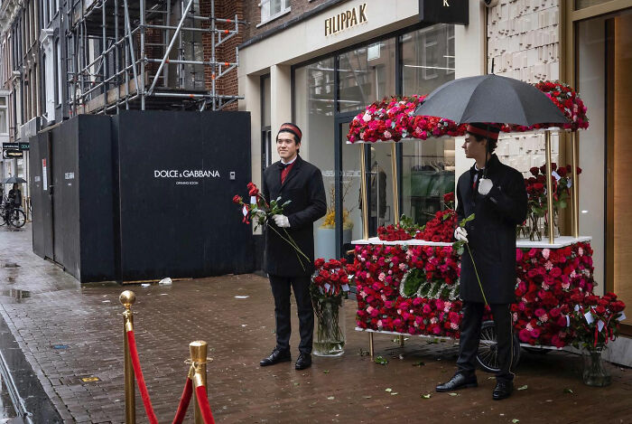 Two men in black suits holding roses stand by a red flower cart and umbrella in a funny accidental street photo.