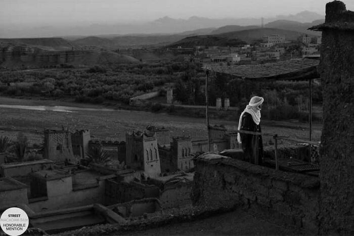 Black and white street photo of a person overlooking traditional buildings in a rural landscape at dusk.