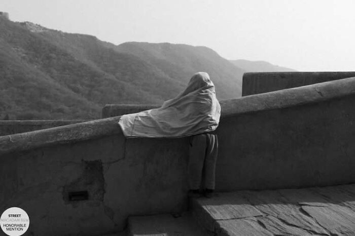 Black and white street photo of a person draped in cloth leaning on a stone railing with mountains in the background.