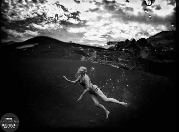 Black and white street photo of a woman swimming underwater with a dramatic cloudy sky above the water surface.