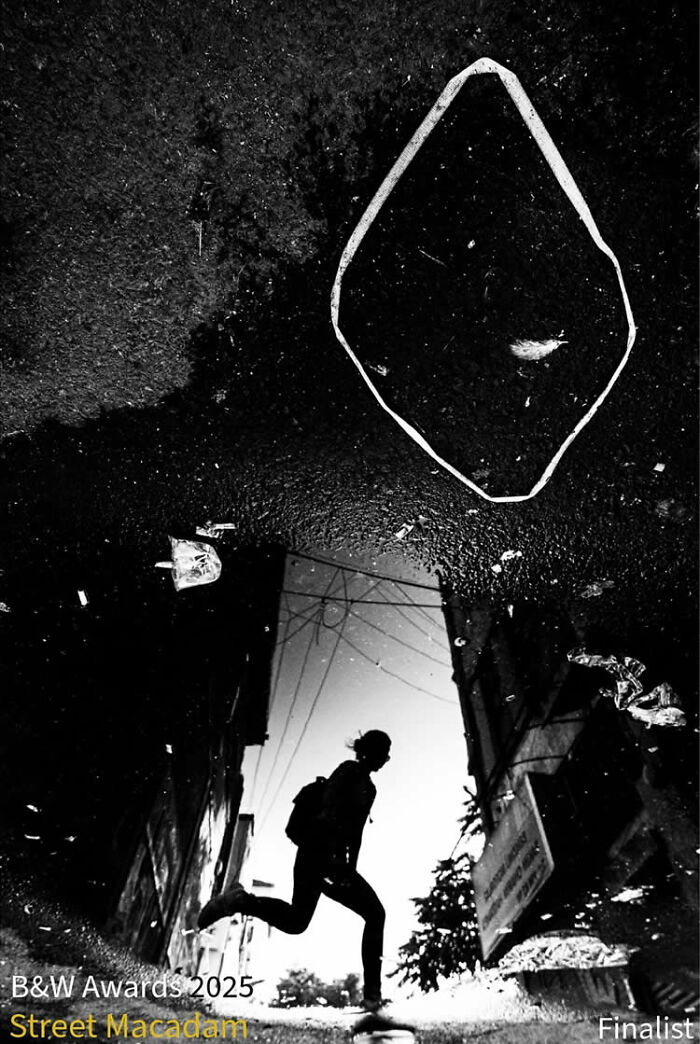 Black and white street photo of a person running reflected in a puddle, capturing urban life for Macadam Awards.