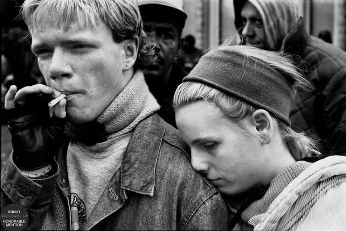 Black and white street photo of a young man smoking with a woman resting her head on his shoulder, capturing raw emotion.