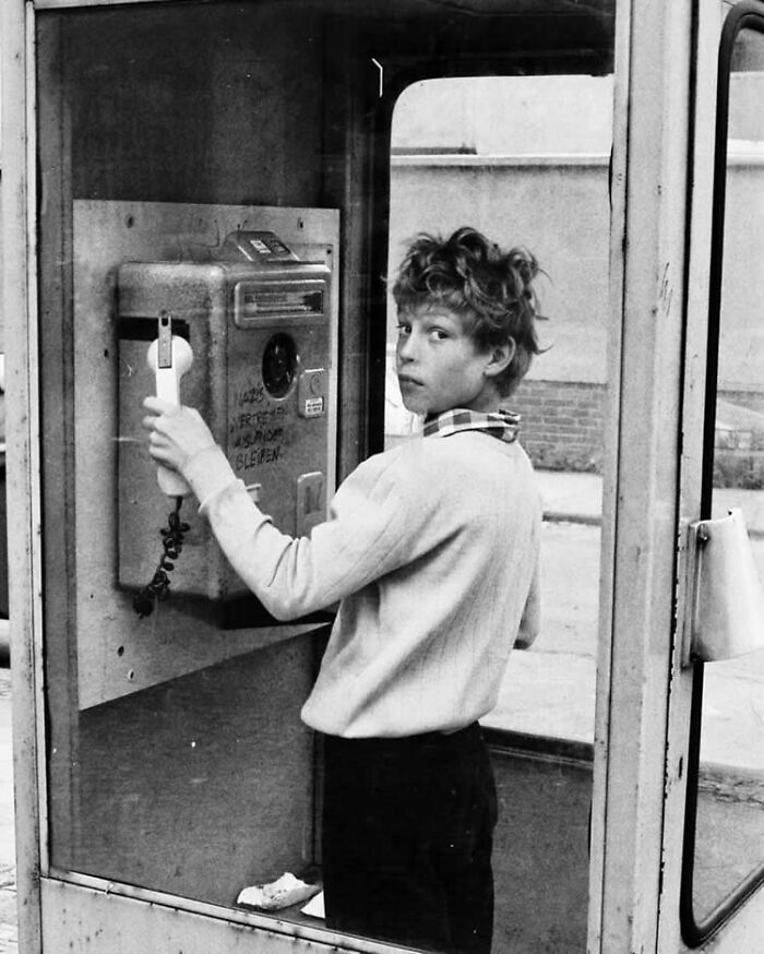 Black and white street photo of a young person using a payphone inside a phone booth, capturing candid urban life.