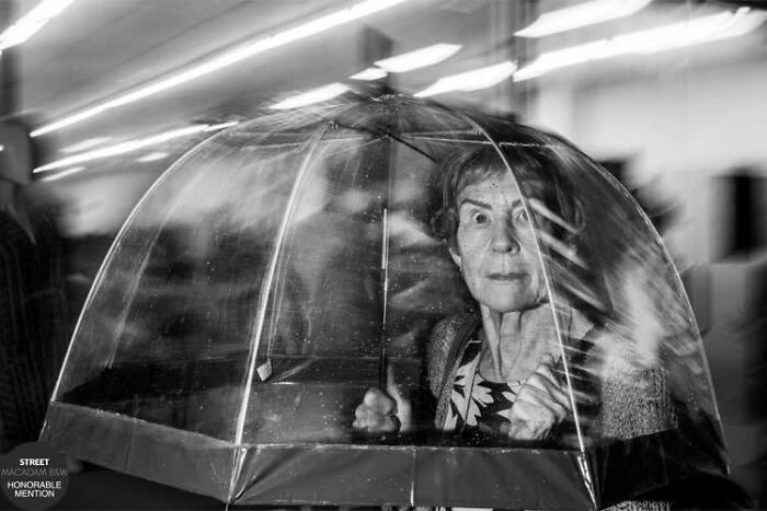 Elderly woman holding a transparent umbrella in a black and white street photo capturing motion and emotion.