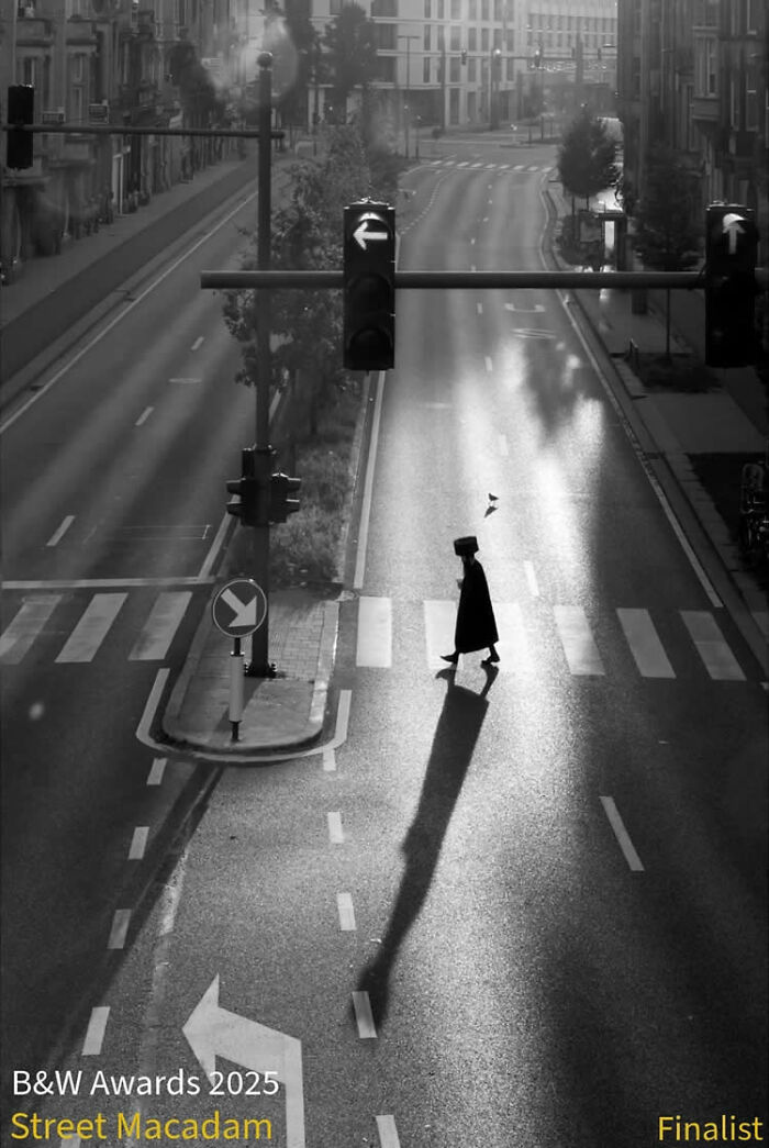 Black and white street photo of a lone pedestrian crossing an empty city intersection at dawn with long shadows.