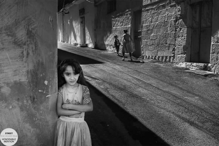 Black and white street photo of a girl leaning against a wall with two children walking on a sunlit road behind her.
