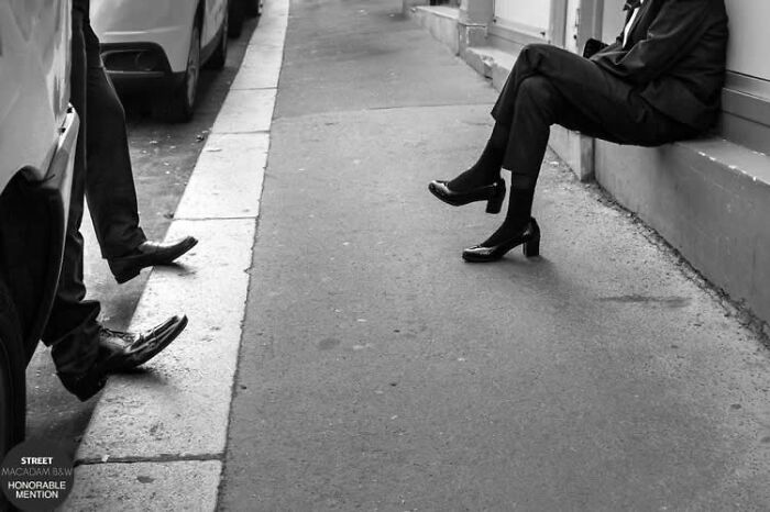 Black and white street photo showing people sitting and standing on a city sidewalk in elegant shoes and attire.