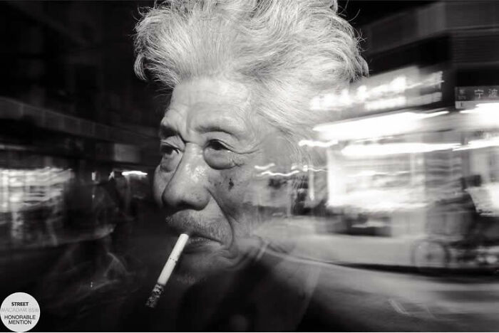 Black and white street photo of an elderly man smoking a cigarette with blurred urban background, Macadam Awards winner.
