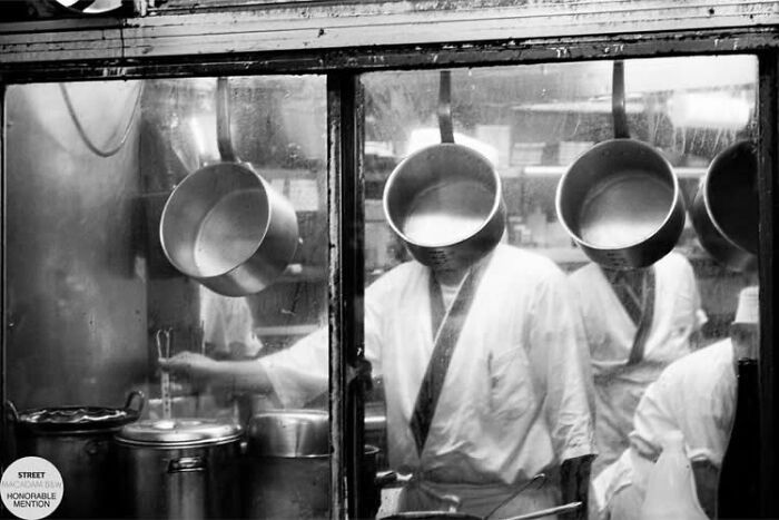Black and white street photo of chefs working in a busy kitchen with pots hanging and steam rising.
