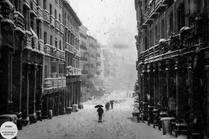 Black and white street photo showing people walking with umbrellas on a snowy urban street, capturing award-winning street photography.